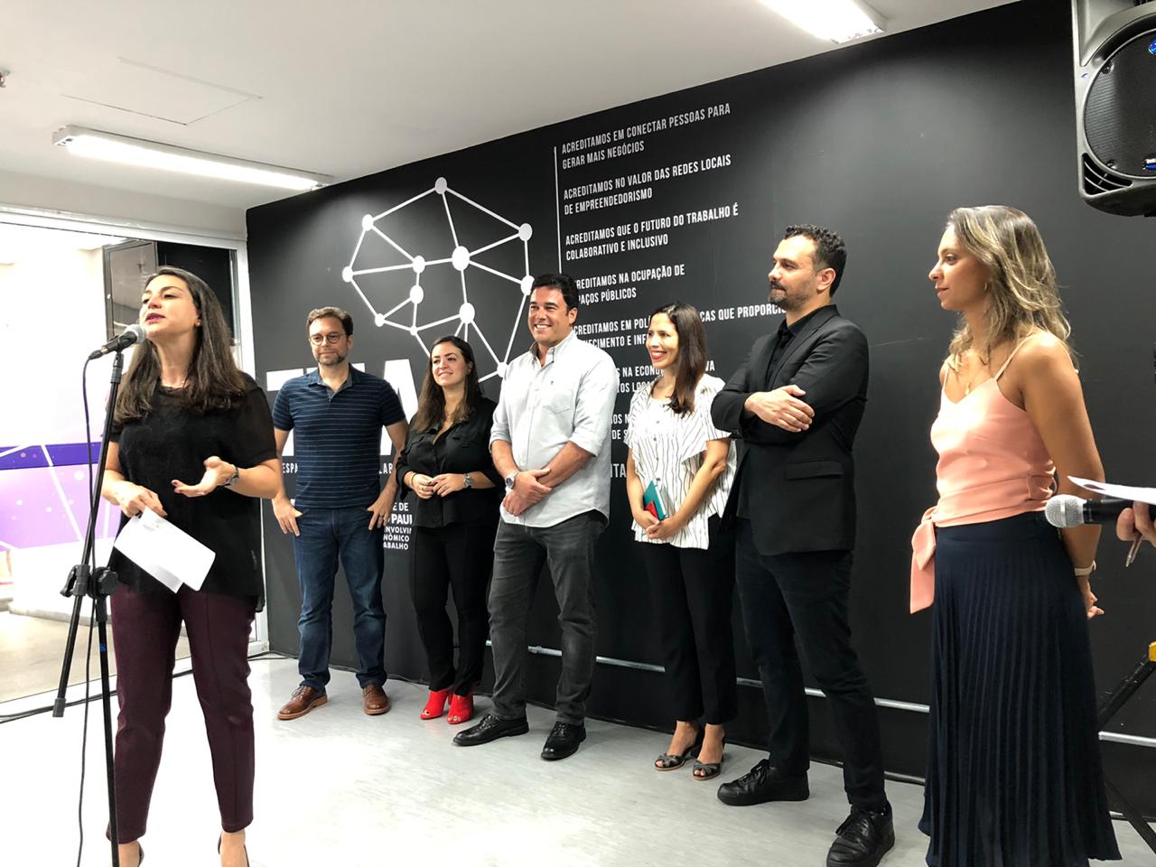 A Secretária de Desenvolvimento Econômico fala ao microfone enquanto os Secretários observam de fundo